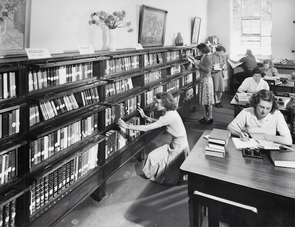 Black and white image of the shelves of a library in the 1930s with women browsing for books and sitting at tables reading