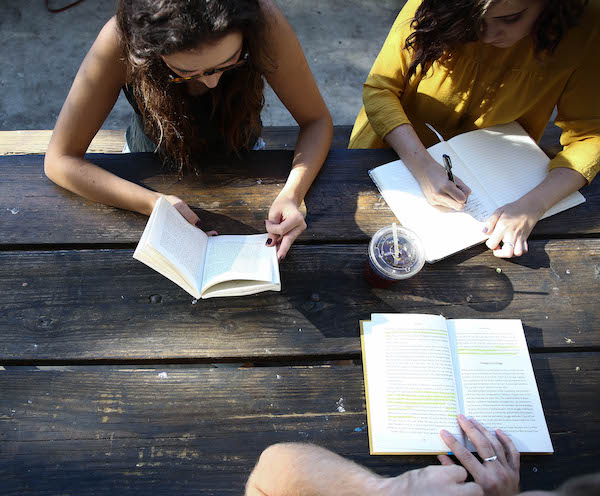 Overhead view of three people sitting outside around a wood table reading.