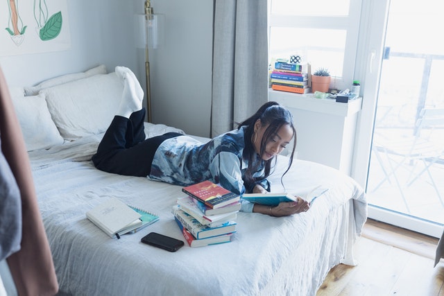 a woman reading on a bed next to a stack of books and notebooks