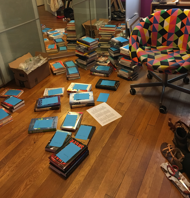 a photo of stacks of books taking over the floor of a room that also includes a swivel chair, various shoes, and a box of packing material